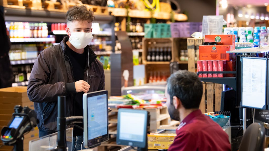 Ein junger Mann trägt bei seinem Einkauf in einem Supermarkt einen Mundschutz, der Verkäufer sitzt hinter einer Plexiglasscheibe.&nbsp;Edeka hat in einigen Filialen nun spezielle eBons an den Kassen eingeführt.&nbsp;