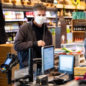 Ein junger Mann trägt bei seinem Einkauf in einem Supermarkt einen Mundschutz, der Verkäufer sitzt hinter einer Plexiglasscheibe. Edeka hat in einigen Filialen nun spezielle eBons an den Kassen eingeführt.