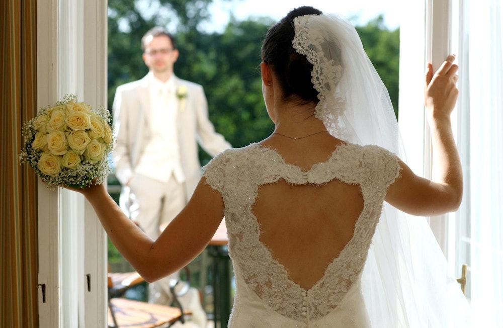 Ein frisch vermähltes Hochzeitspaar. Sie trägt Hochzeitskleid und Brautstrauß und schaut ihn durch Terrassentür an.
