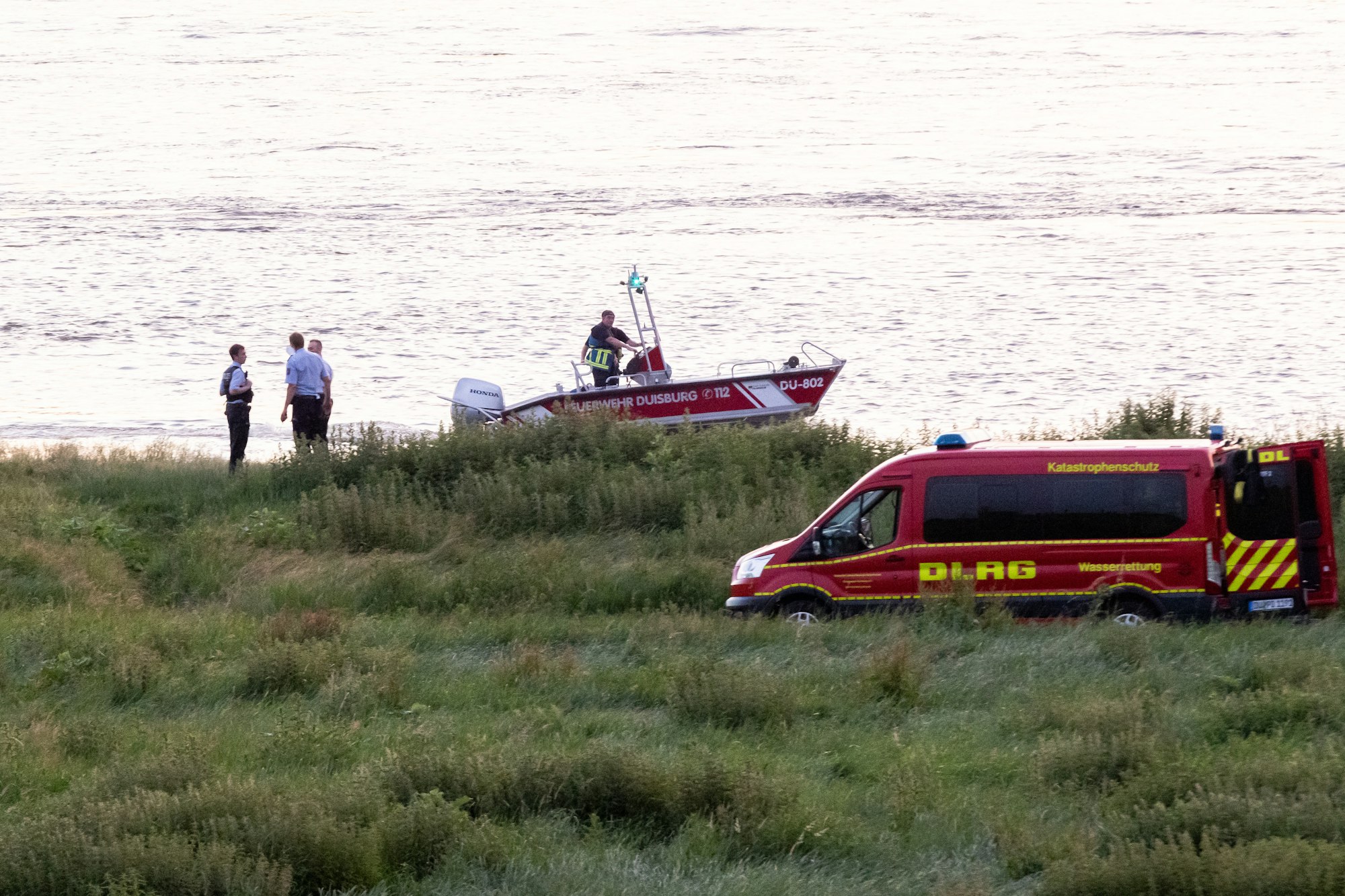 Einsatzkräfte der Polizei, Feuerwehr und DLRG arbeiten am Ufer des Rheins. Auch ein Rettungsboot ist im Einsatz.