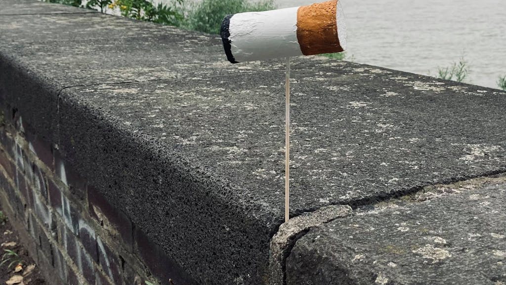 Eine überdimensionale Zigarettenkippe ist auf einer Mauer am Kölner Rheinufer befestigt, im Hintergrund der Dom.&nbsp;