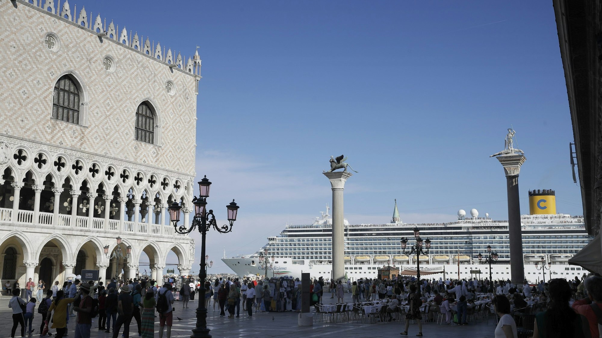 Ein Kreuzfahrtschiff fährt im Sommer 2019 am Markusplatz in Venedig vorbei, auf dem Platz stehen zahlreiche Menschen, die Sonne scheint. Italiens Regierung hat ein Durchfahrtsverbot für große Kreuzfahrtschiffe durch einen Teil der Lagune von Venedig beschlossen.