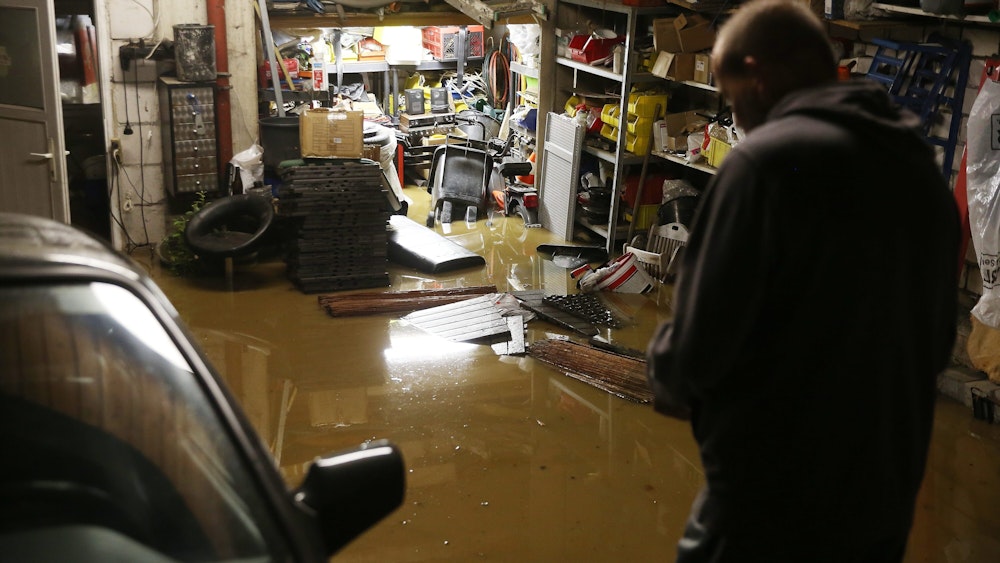 Ein Hausbesitzer steht in seiner überfluteten Garage, Regale stehen unter Wasser, ein Auto steht neben ihm.