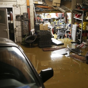 Ein Hausbesitzer steht in seiner überfluteten Garage, Regale stehen unter Wasser, ein Auto steht neben ihm.