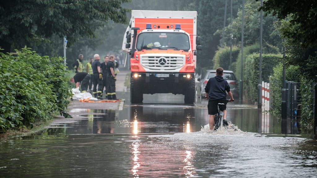 Ein Fahrradfahrer fährt am 14. Juli 2021 durch eine überflutete Straße in der Düsseldorfer Ostparksiedlung. Im Hintergrund steht ein Einsatzfahrzeug der Rettungskräfte.