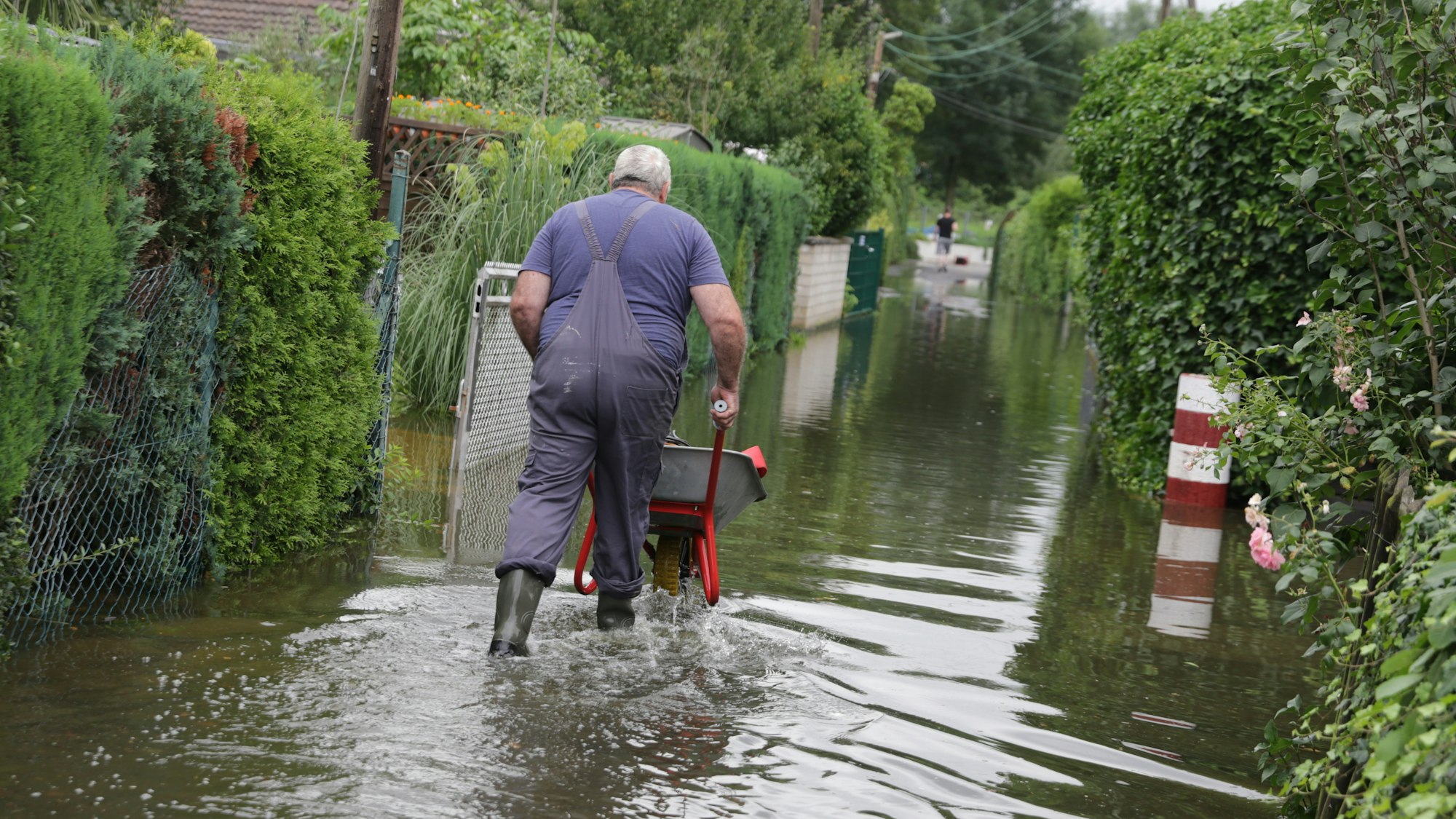 Ein Mann schiebt eine Schubkarre durch das knöcheltiefe Wasser eines überschwemmten Weges.