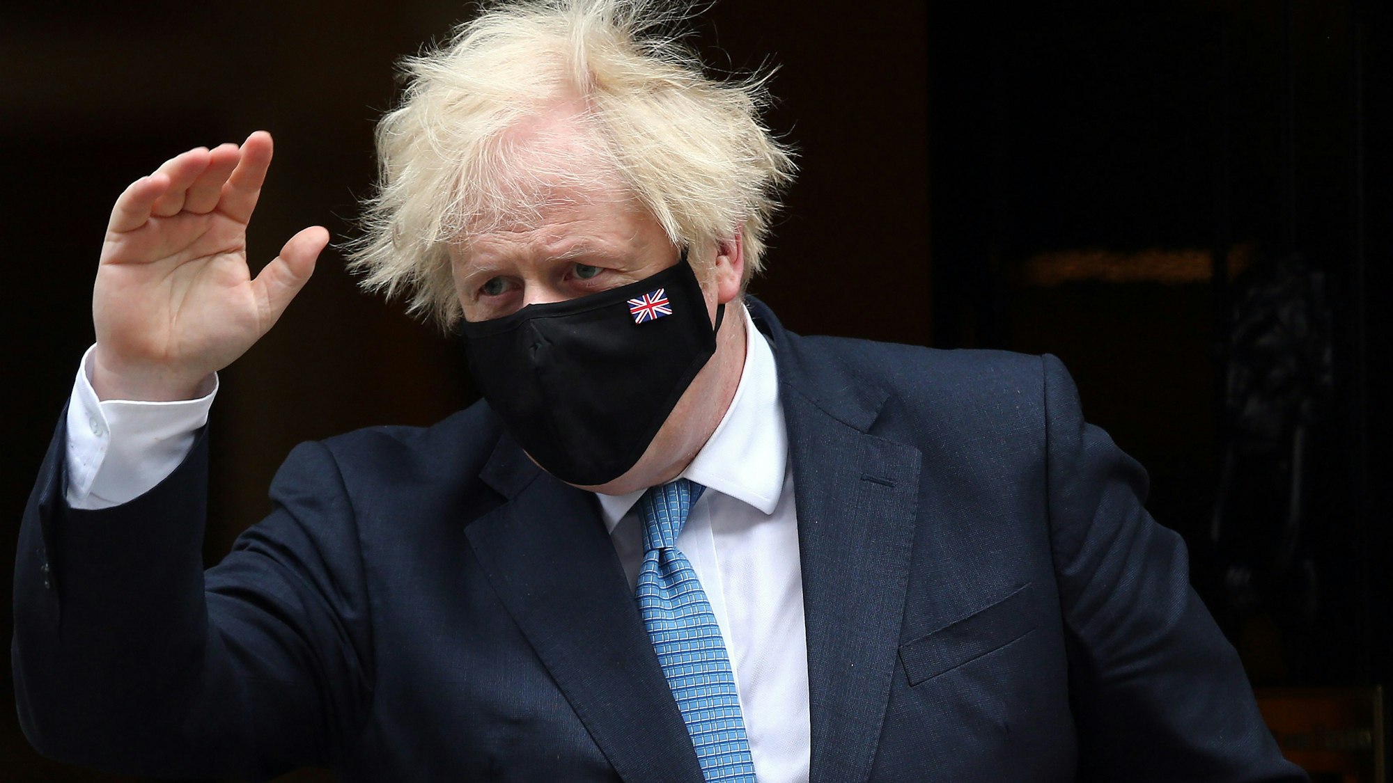 Großbritanniens Premier Minister Boris Johnson trägt eine dunkle Maske und winkt mit der rechten Hand.
