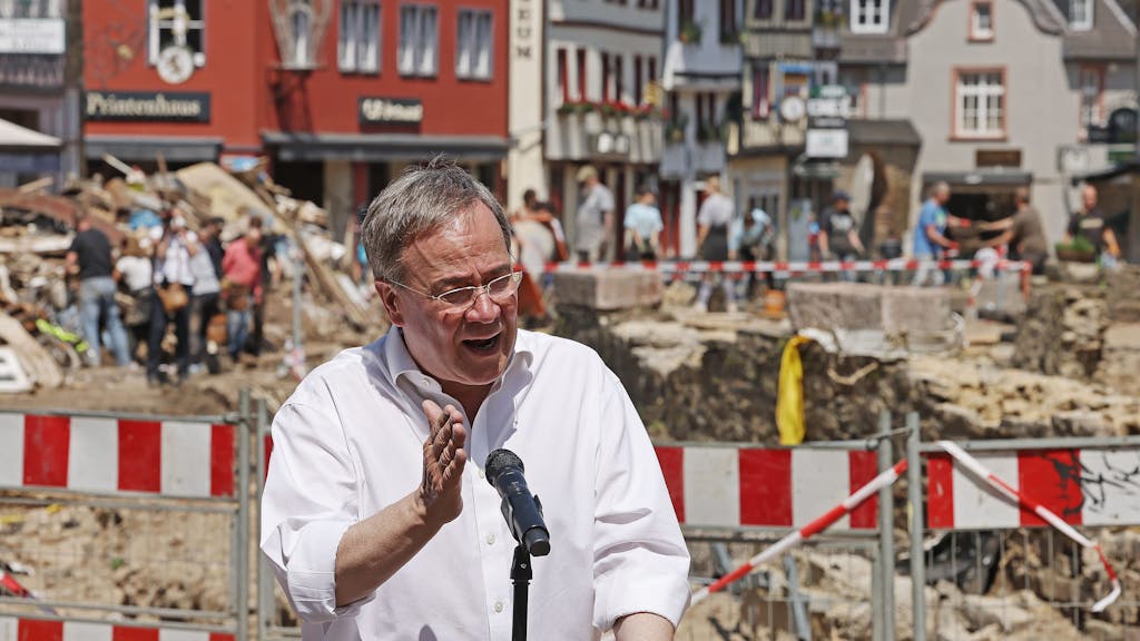 Armin Laschet spricht in ein Mikrofon. Im Hintergrund sieht man die Unwetterschäden in Bad Münstereifel, die mit einem Bauzaun abgesperrt sind.