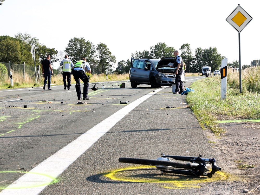 Das abgerissene Vorderrad eines Fahrrades liegt neben der mit Unfalltrümmern übersäten Straße, auf der Polizisten den Unfall aufnehmen. Im Hintergrund steht das am Unfall beteiligte Auto.