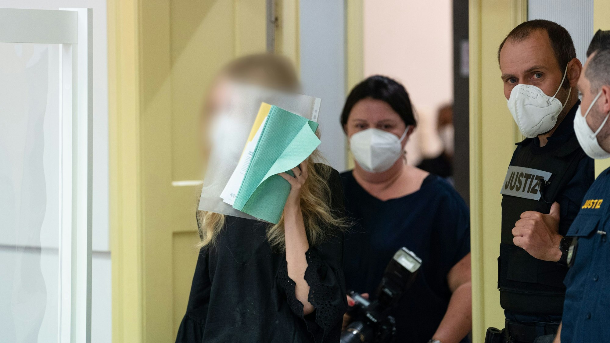 Eine Frau hält sich im Landgericht Schweinfurt Unterlagen vor das Gesicht. Um sie herum stehen Justiz-Beamte mit Masken.