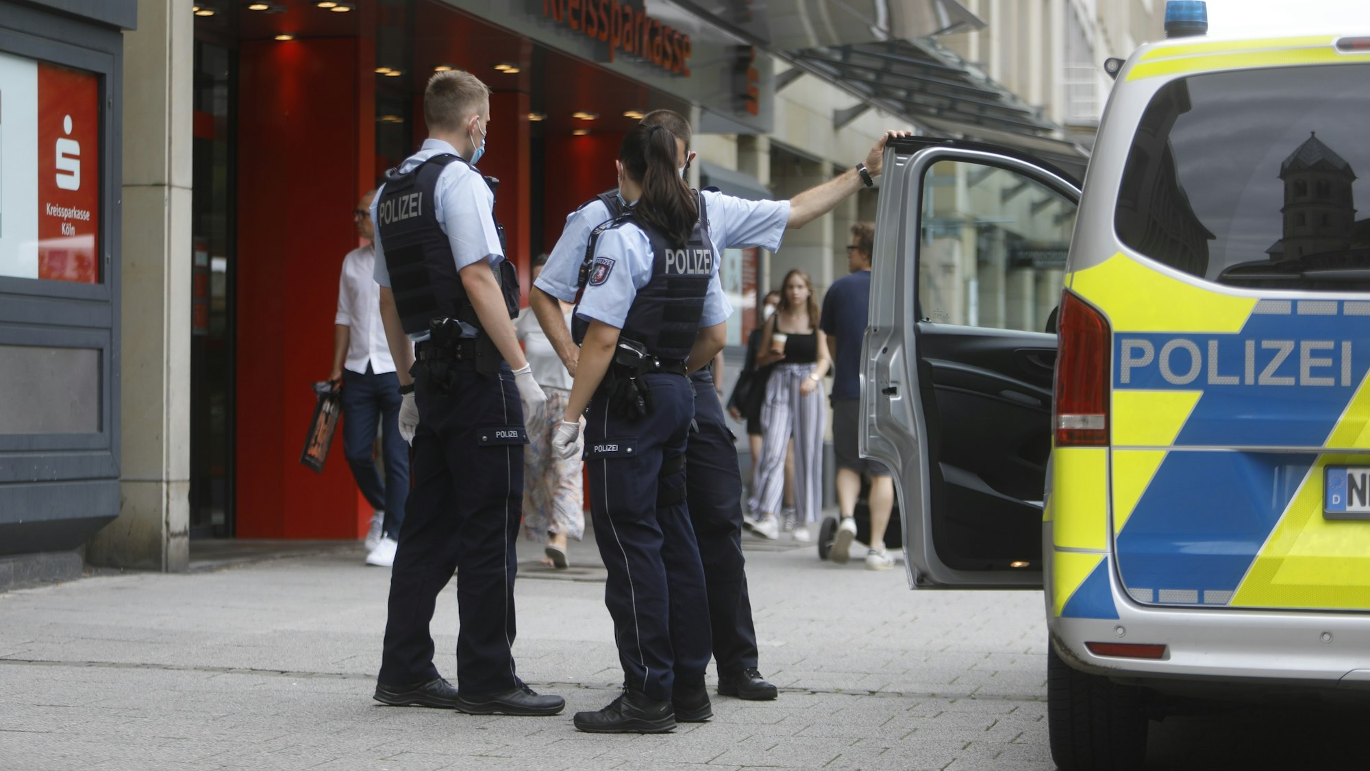 Zwei Polizisten und eine Polizistin stehen neben einem Einsatzfahrzeug vor einer Sparkasse.