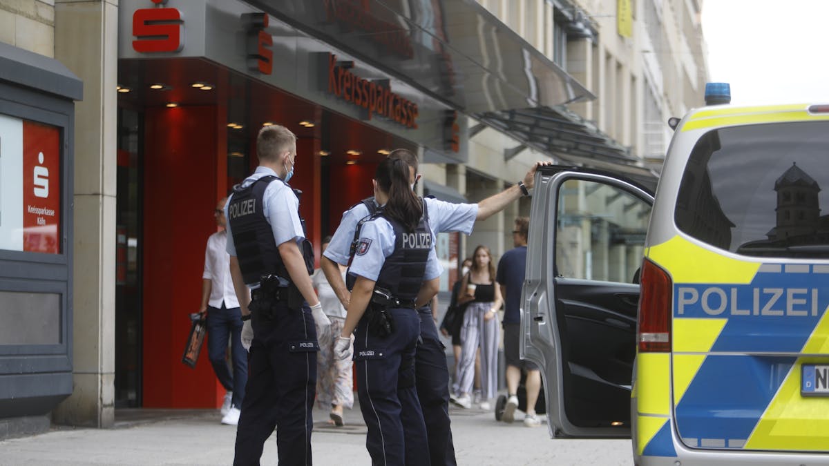 Zwei Polizisten und eine Polizistin stehen neben einem Einsatzfahrzeug vor einer Sparkasse.&nbsp;