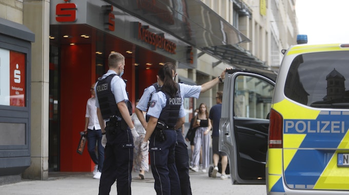 Zwei Polizisten und eine Polizistin stehen neben einem Einsatzfahrzeug vor einer Sparkasse.