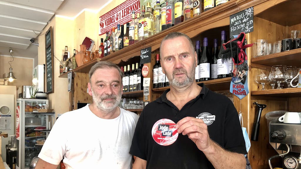 Rüdiger Nehl und Dieter Niehoff stehen hinter der Theke im Restaurant und halten den „Jode Lade“-Sticker in die Kamera.
