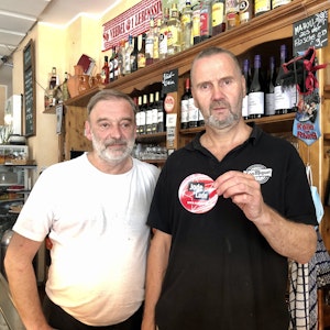 Rüdiger Nehl und Dieter Niehoff stehen hinter der Theke im Restaurant und halten den „Jode Lade“-Sticker in die Kamera.