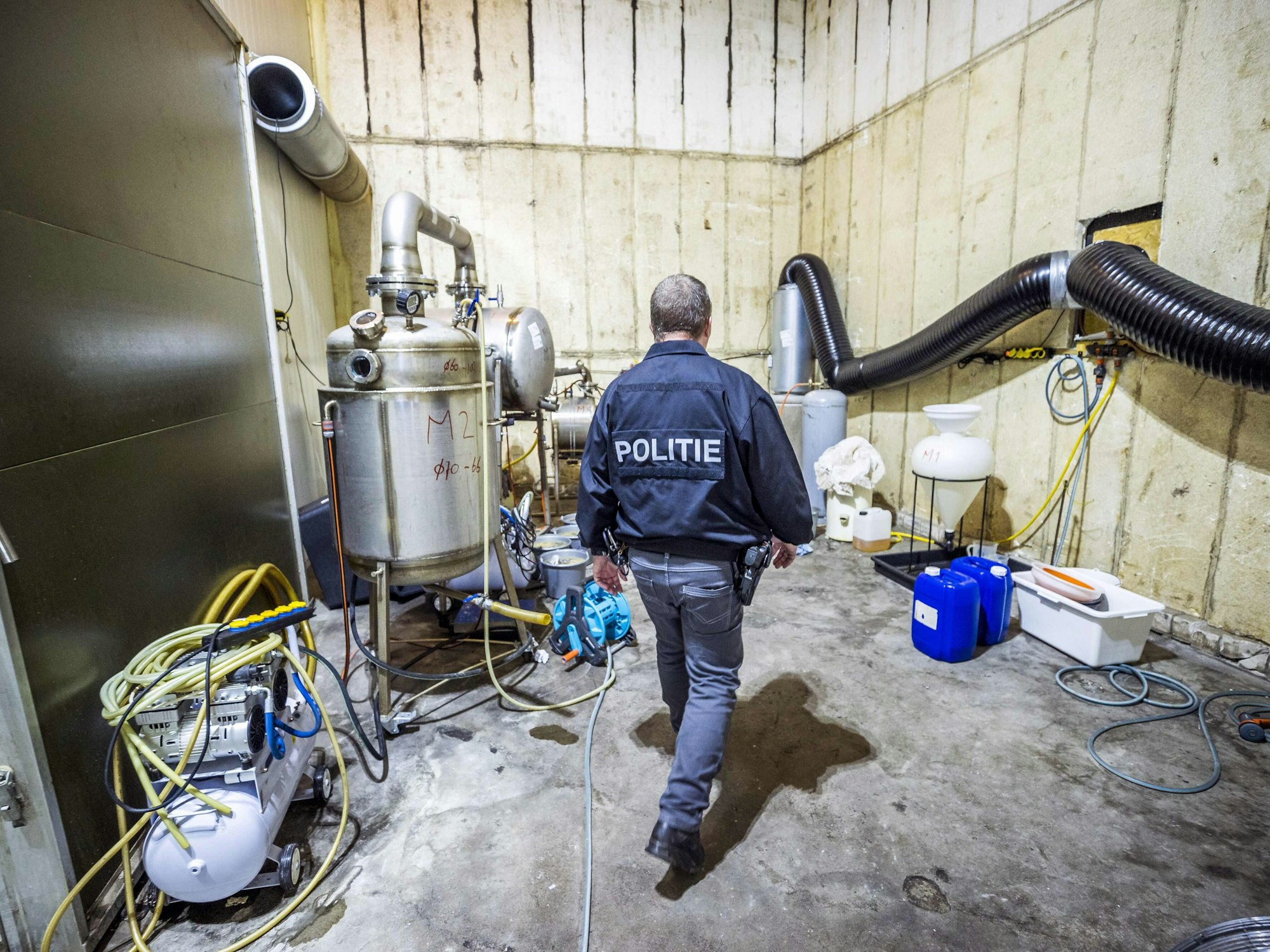 In einem Lagerraum stehen Apparate zur Herstellung chemischer Drogen. Ein Polizei-Beamter läuft durch das Labor.