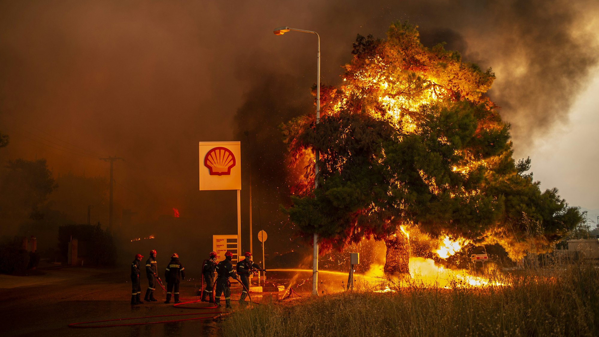 Feuerwehrleute bekämpfen einen Waldbrand in einem Waldgebiet nördlich von Athen, welches sich einer Tankstelle nähert.