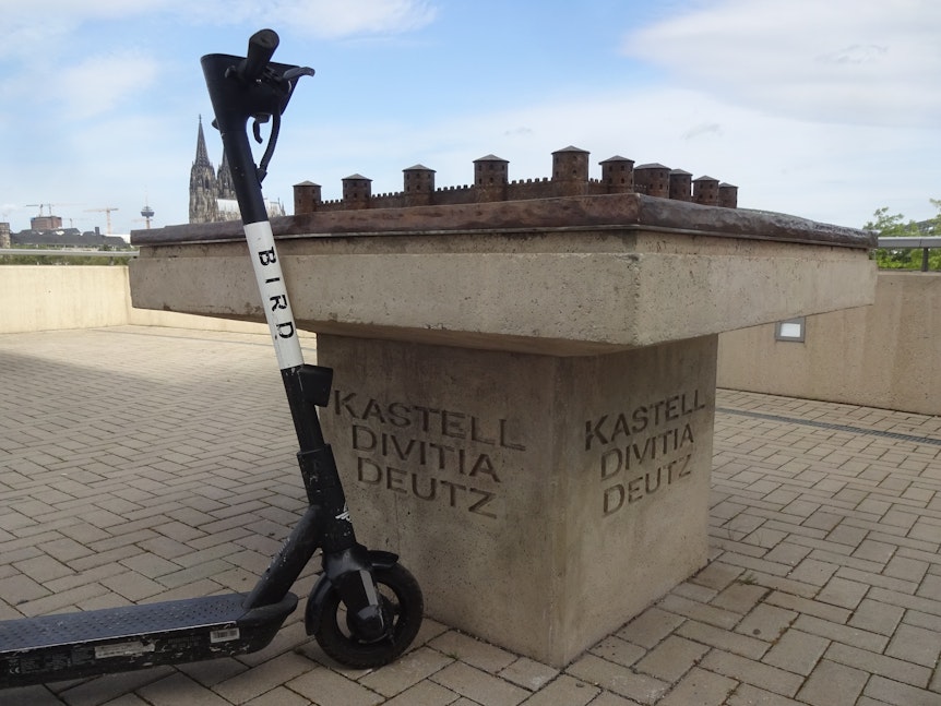 Ein E-Scooter steht neben dem Bronzemodell des Deutzer Kastells in Köln.