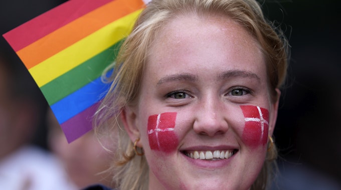 Eine Frau hat sich die Dänemark-Flagge auf ihre Wangen gemalt. Im Hintergrund sieht man eine Regenbogen-Fahne, das Zeichen der LGBTQI-Community.