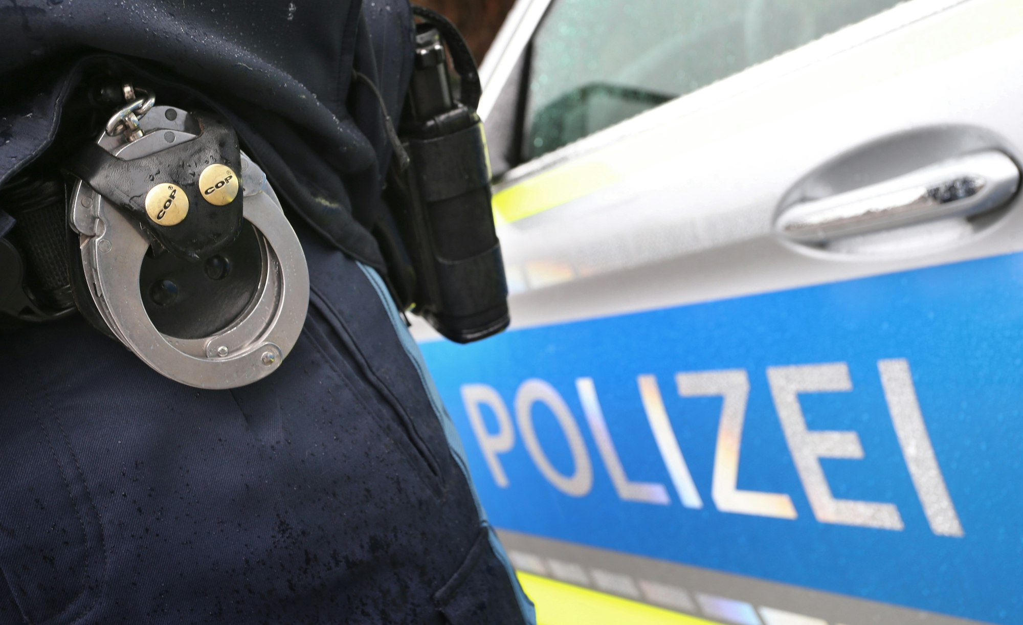Ein Polizist mit Handschellen und einer Pistole am Gürtel steht vor einem Streifenwagen, aufgenommen im März 2019.