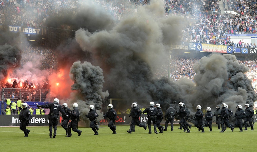 HSV Ultras zünden Pyro und Rauchbomben. Das Stadion ist bedeckt von einer Rauchwolke, die Polizei läuft auf den Rasen.