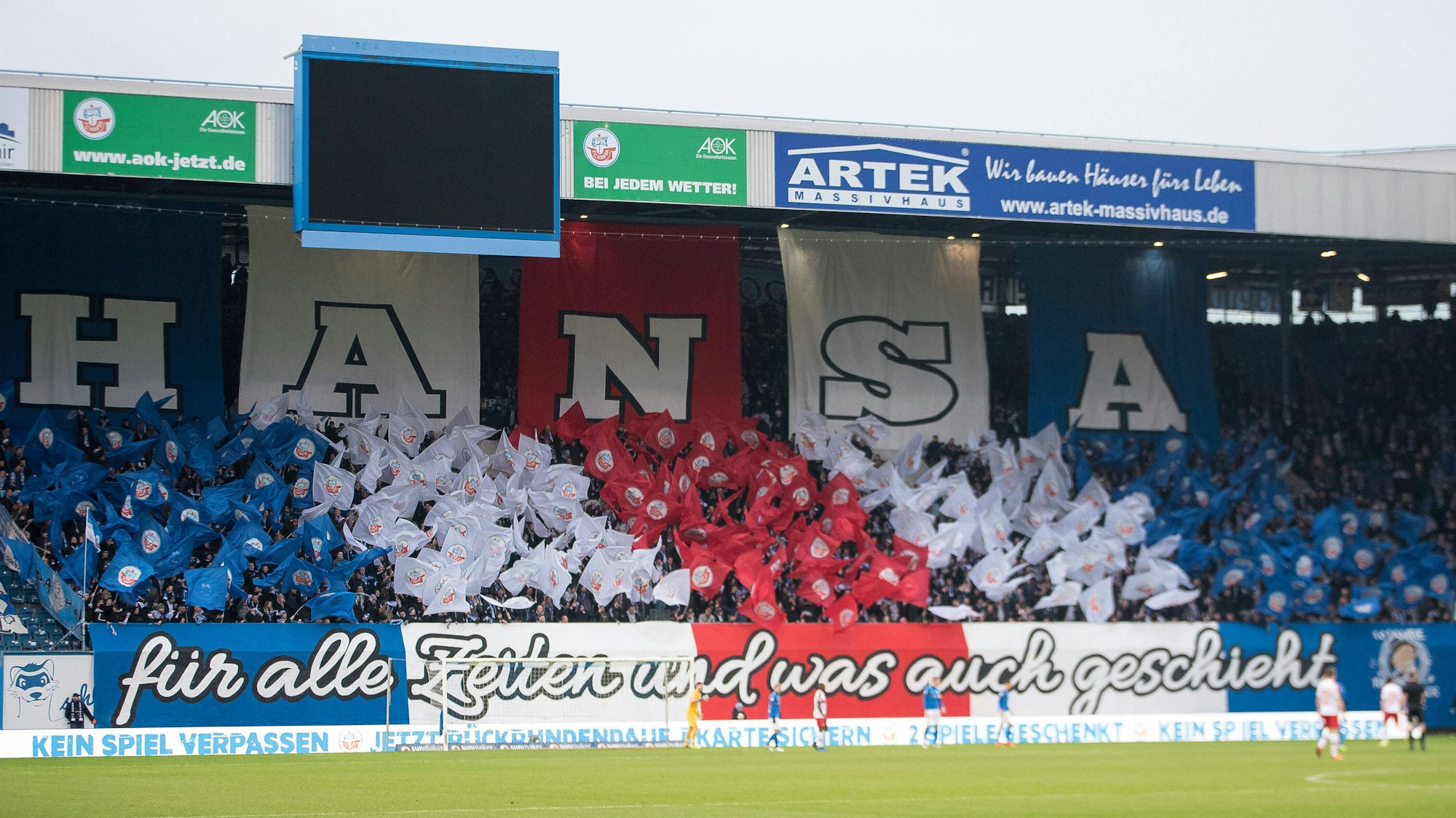 Die Kurve des Hansa-Fans im Ostseestadion ist bedeckt mit blauen, roten und weißen Fahnen und einem Schriftzug: „Hansa – für alle Zeiten und was auch geschieht“.
