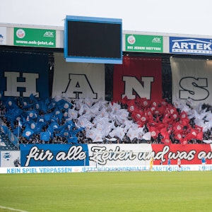 Die Kurve des Hansa-Fans im Ostseestadion ist bedeckt mit blauen, roten und weißen Fahnen und einem Schriftzug: „Hansa – für alle Zeiten und was auch geschieht“.