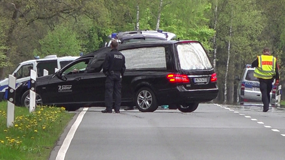 Ein Leichenwagen steht auf einer Straße, davor ein Polizist.