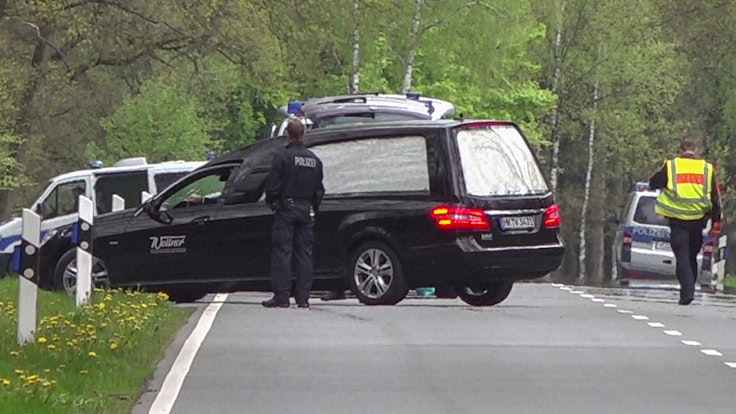 Ein Leichenwagen steht auf einer Straße, davor ein Polizist.