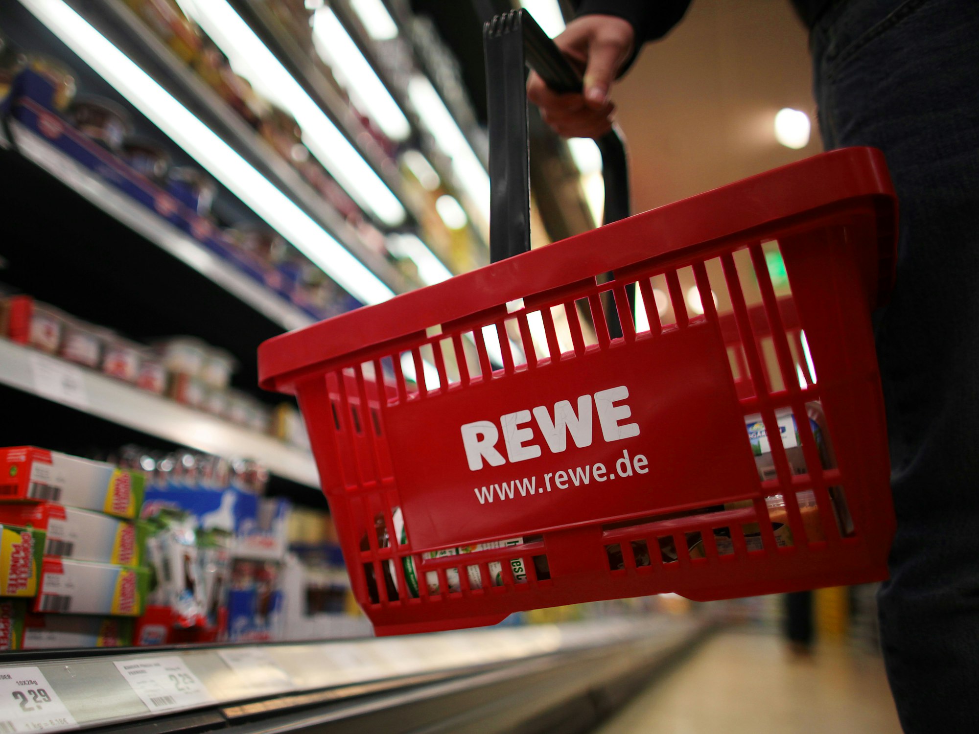 Ein Kunde geht in einem Rewe-Supermarkt mit einem Einkaufskorb am Kühlregal entlang.