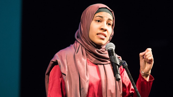 Nemi El-Hassan steht in Braunschweig in einem roten Kleid und mit rotem Kopftuch am Mikrofon auf einer Bühne bei einem Poetry Slam.