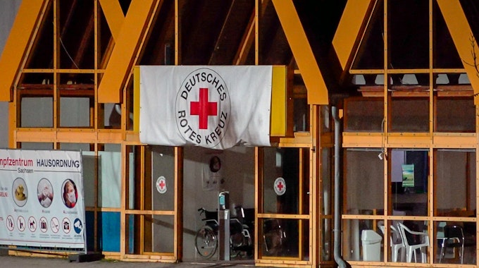 Eine Rot-Kreuz-Fahne hängt über dem Eingang zum Impfzentrum in Eich. Im sächsischen Vogtlandkreis hat es einen Brandanschlag auf ein Impfzentrum gegeben.