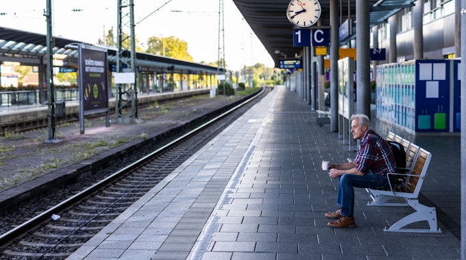 Ein Mann sitzt mit einem Kaffee in der Hand auf einer Bank auf dem ansonsten menschenleeren Bahnsteig.