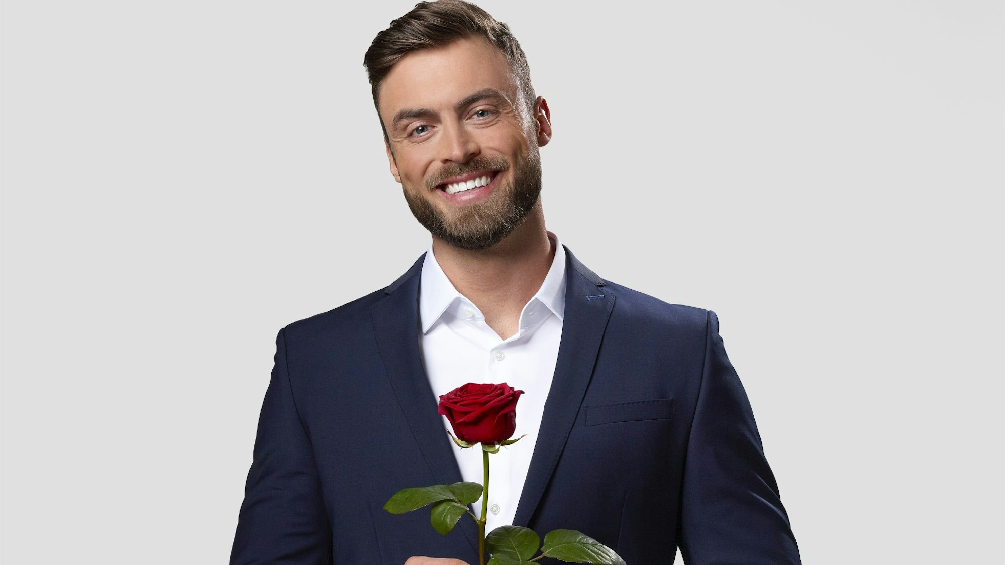 Niko Griesert steht im blauen Anzug und mit einer roten Rose in der Hand vor einer weißen Fotowand.