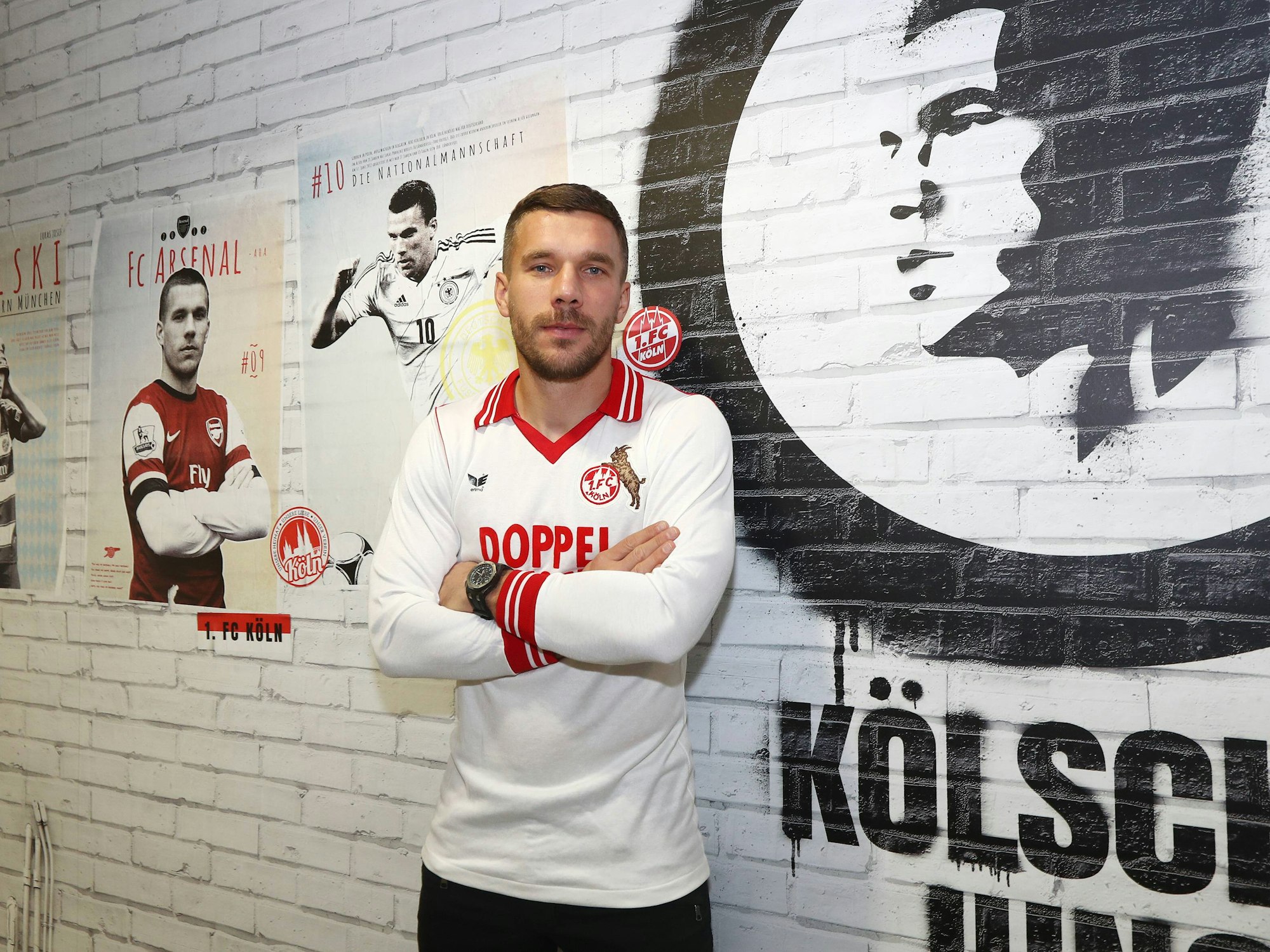 Podolski blickt in die Kamera, verschränkt die Arme. Er trägt ein Traditionstrikot des 1. FC Köln.