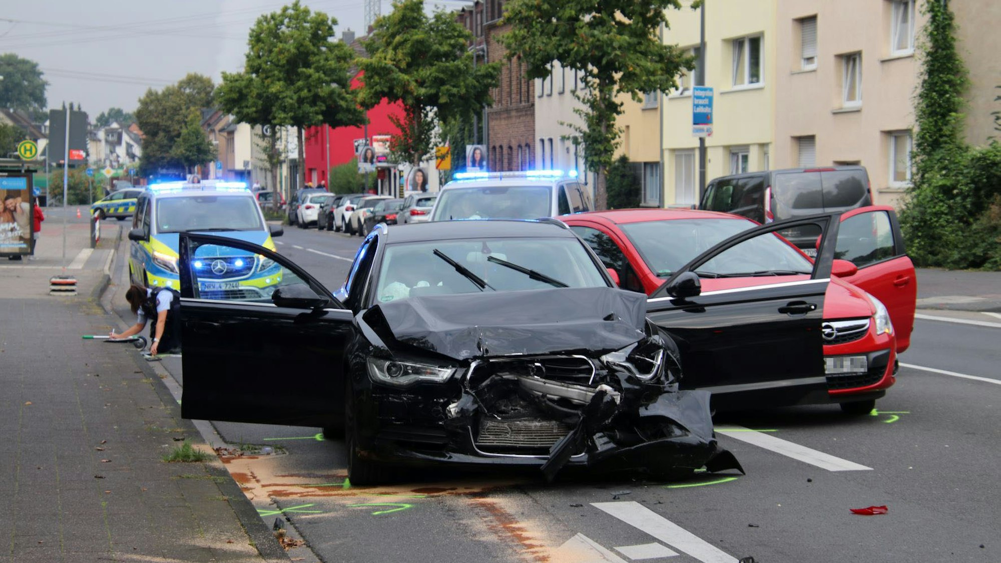 Ein Auto mit demolierter Front steht neben einem anderen Unfallfahrzeug, im Hintergrund steht die Polizei mit Blaulicht auf der Straße.