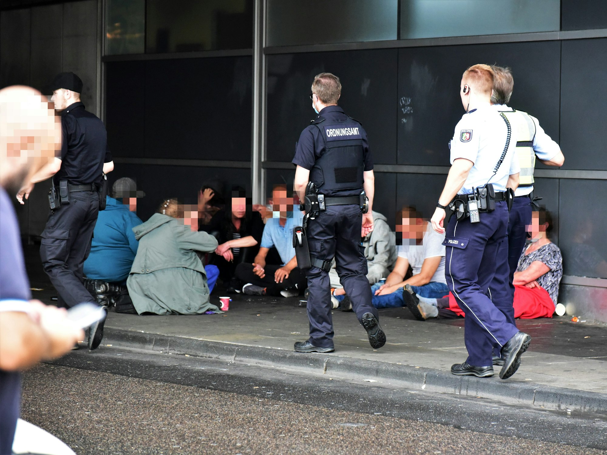 Mehrere Menschen, offenbar aus der Trinker- und Drogenszene, sitzen auf dem Bürgersteig, Polizei und Ordnungsamt gehen an ihnen vorbei.