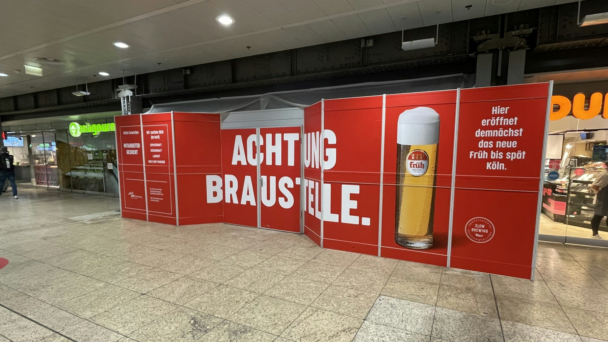 Ein Bauschild mit der Aufschrift „Achtung Braustelle“ steht im Kölner Hauptbahnhof und kündigt das neue Lokal der Brauerei Früh an.