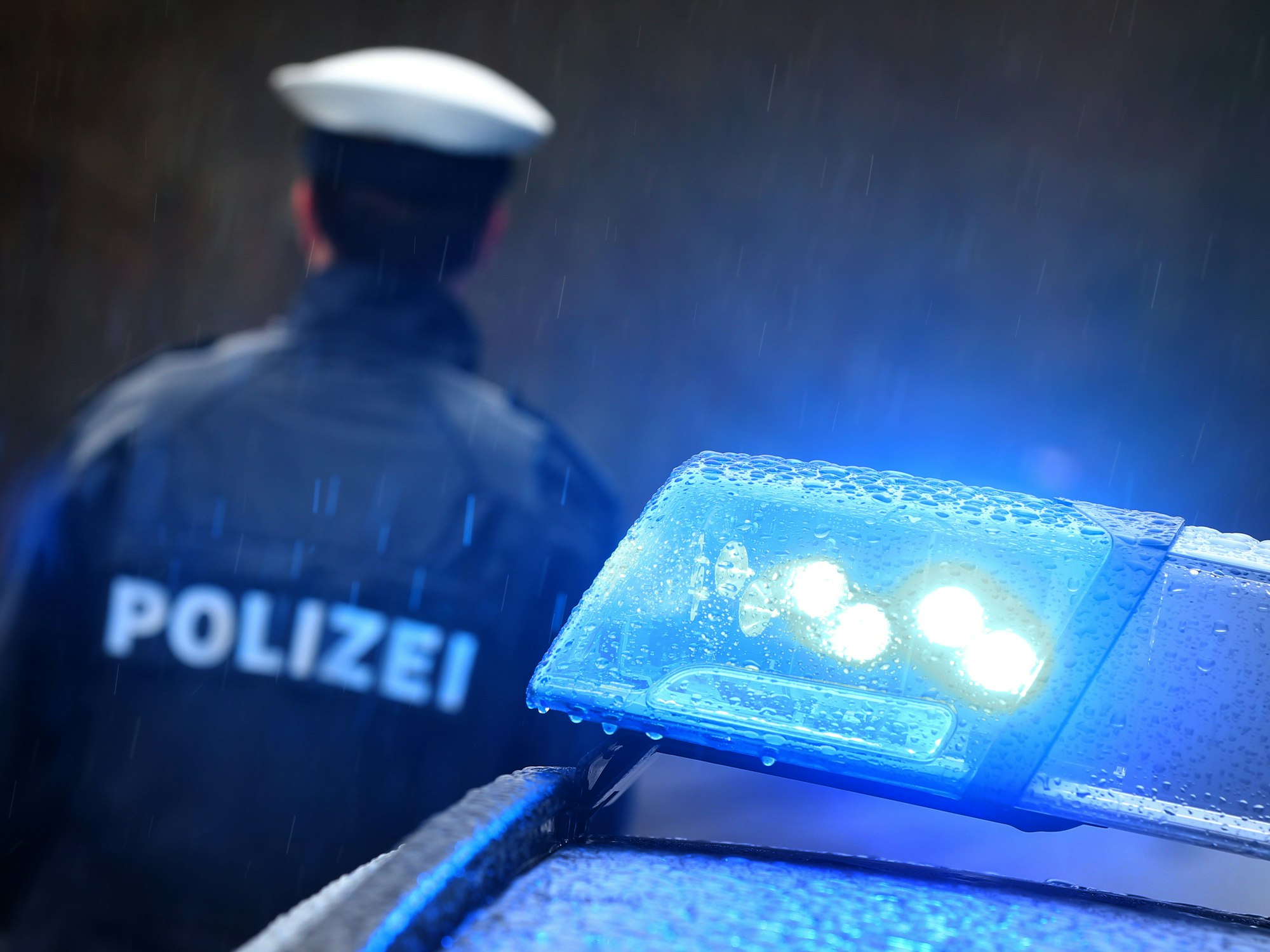 Oberpfalz: Ein Polizist steht im Regen vor einem Streifenwagen dessen Blaulicht aktiviert ist.