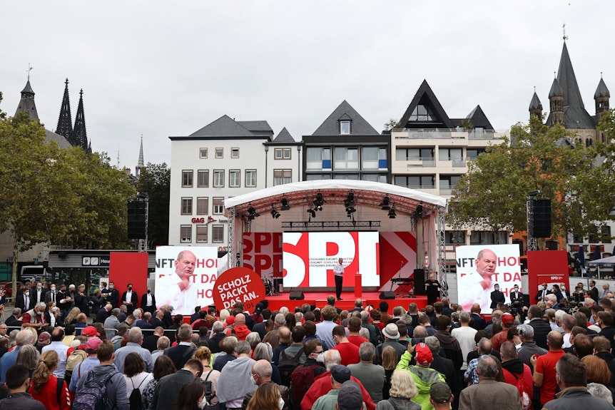 Menschen stehen vor einer Bühne, auf der SPD-Kanzlerkandidat Olaf Scholz spricht.