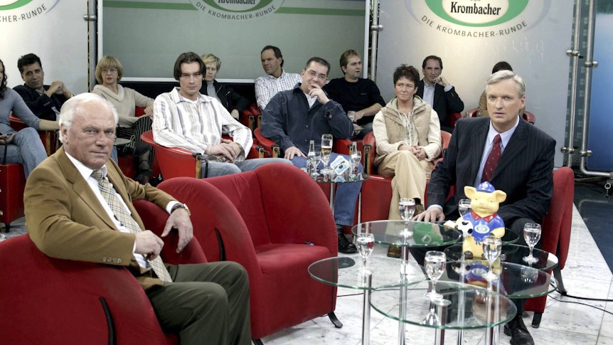 Udo Lattek sitzt links, Rudi Brückner rechts. Zwischen den beiden steht ein freier Sessel. Im Hintergrund sitzen mehrere Zuschauer.