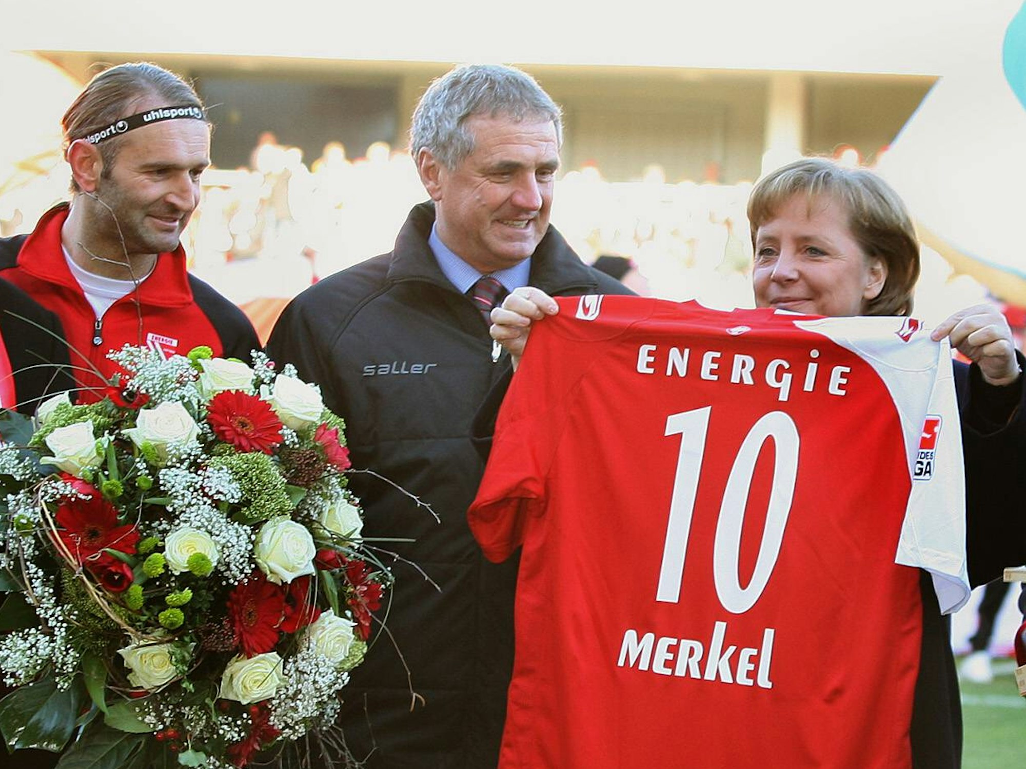 Angela Merkel hält ein Cottbus-Trikot mit der Nummer 10 hoch, Tomislav Piplica hält einen Blumenstrauß in den Händen.