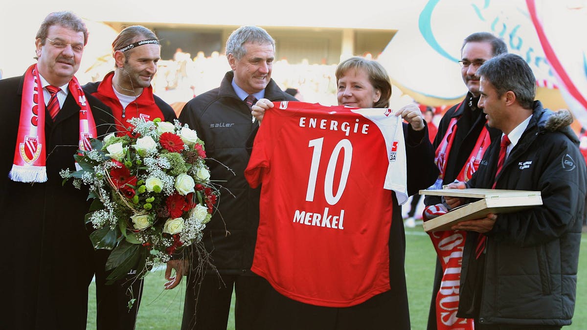 Angela Merkel hält ein Cottbus-Trikot mit der Nummer 10 hoch, Tomislav Piplica hält einen Blumenstrauß in den Händen.