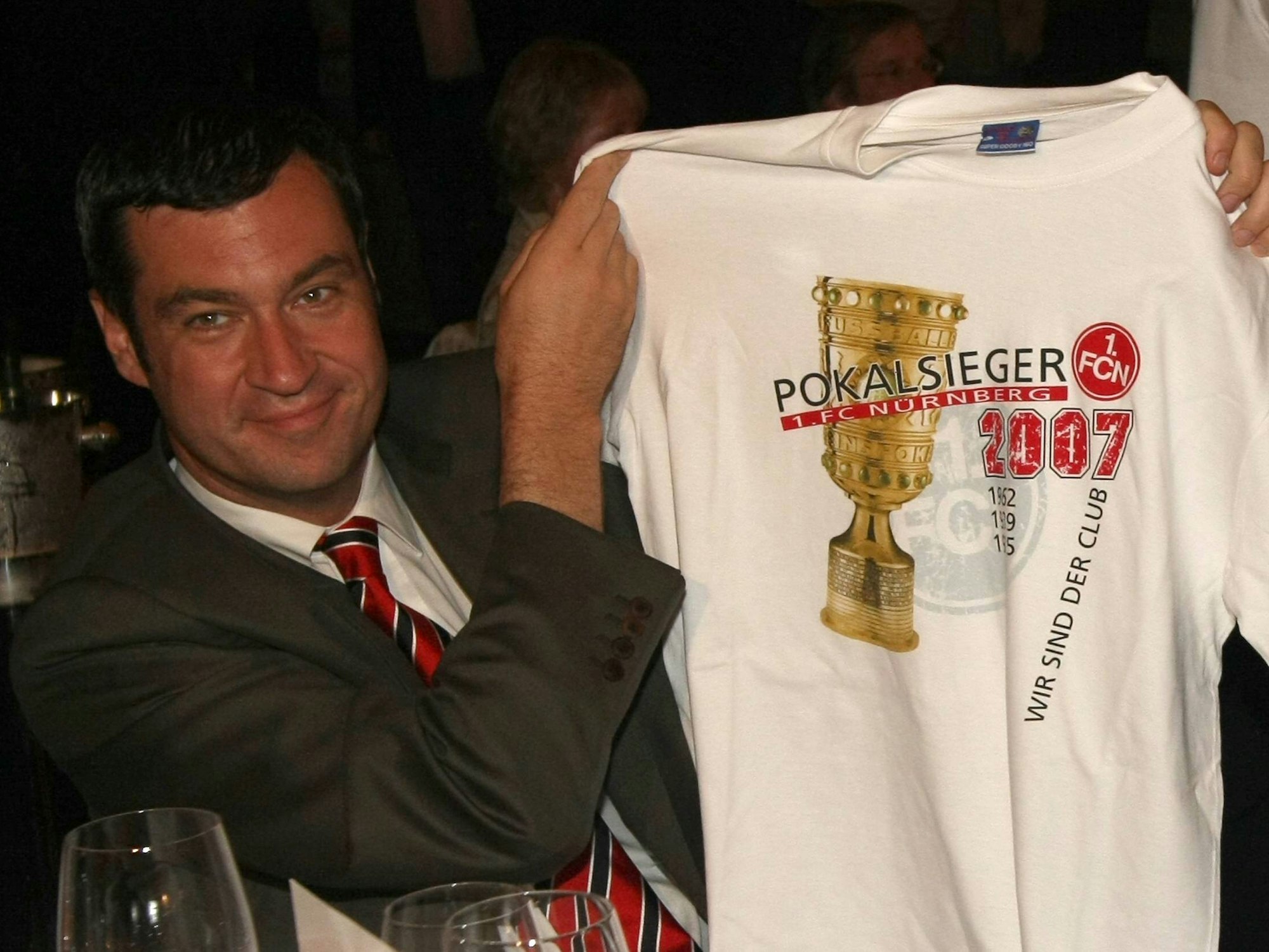 Markus Söder hält ein weißes T-Shirt hoch, auf dem Pokalsieger 2007 steht und der DFB-Pokal abgebildet ist.