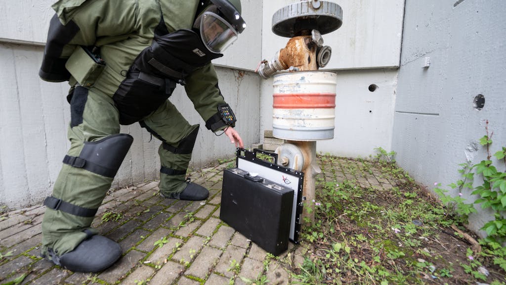Ein Entschärfer im Bombenschutzanzug stellt eine Rötgenplatte für ein mobiles Röntgengerät vor einen Koffer, der an einem Hydranten lehnt.&nbsp;
