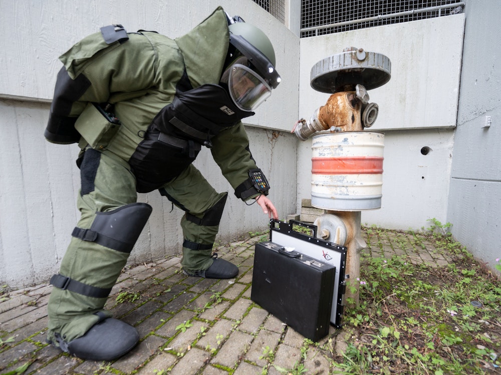Ein Entschärfer im Bombenschutzanzug stellt eine Rötgenplatte für ein mobiles Röntgengerät vor einen Koffer, der an einem Hydranten lehnt.
