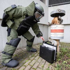 Ein Entschärfer im Bombenschutzanzug stellt eine Rötgenplatte für ein mobiles Röntgengerät vor einen Koffer, der an einem Hydranten lehnt.
