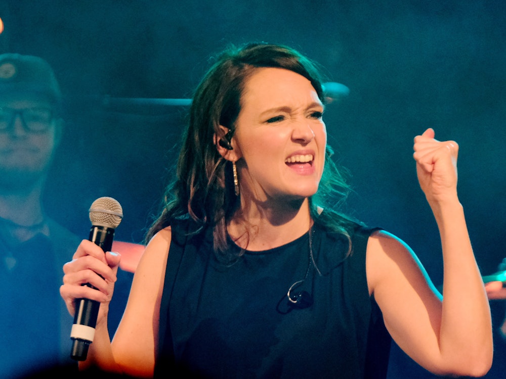 Carolina Kebekus steht mit einem Mikrofon in der Hand auf einer Bühne.