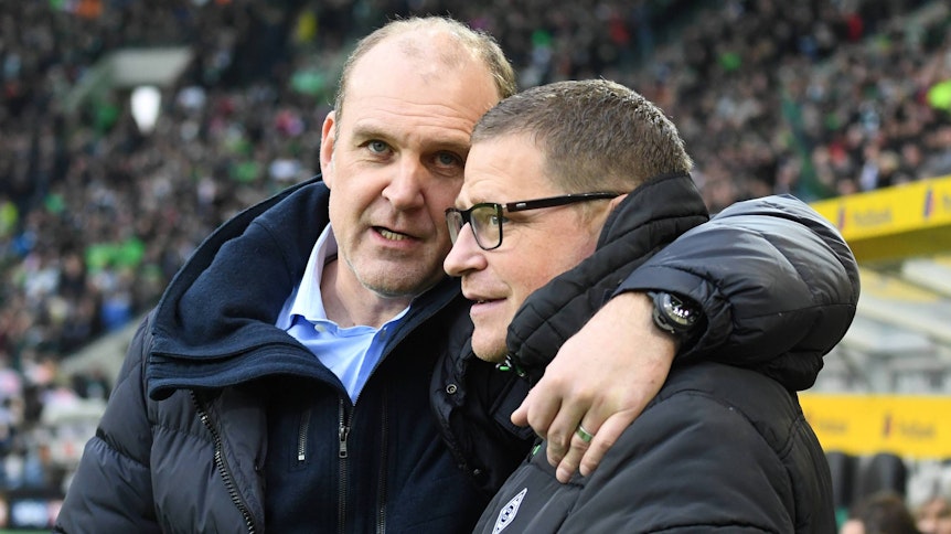 Jörg Schmadtke (l.), damals noch in Diensten des 1. FC Köln, umarmt am 19.11.2016 im Borussia-Park seinen Kumpel und Gladbach-Manager Max Eberl (r.) und schaut dabei in die Kamera eines Fotografen.