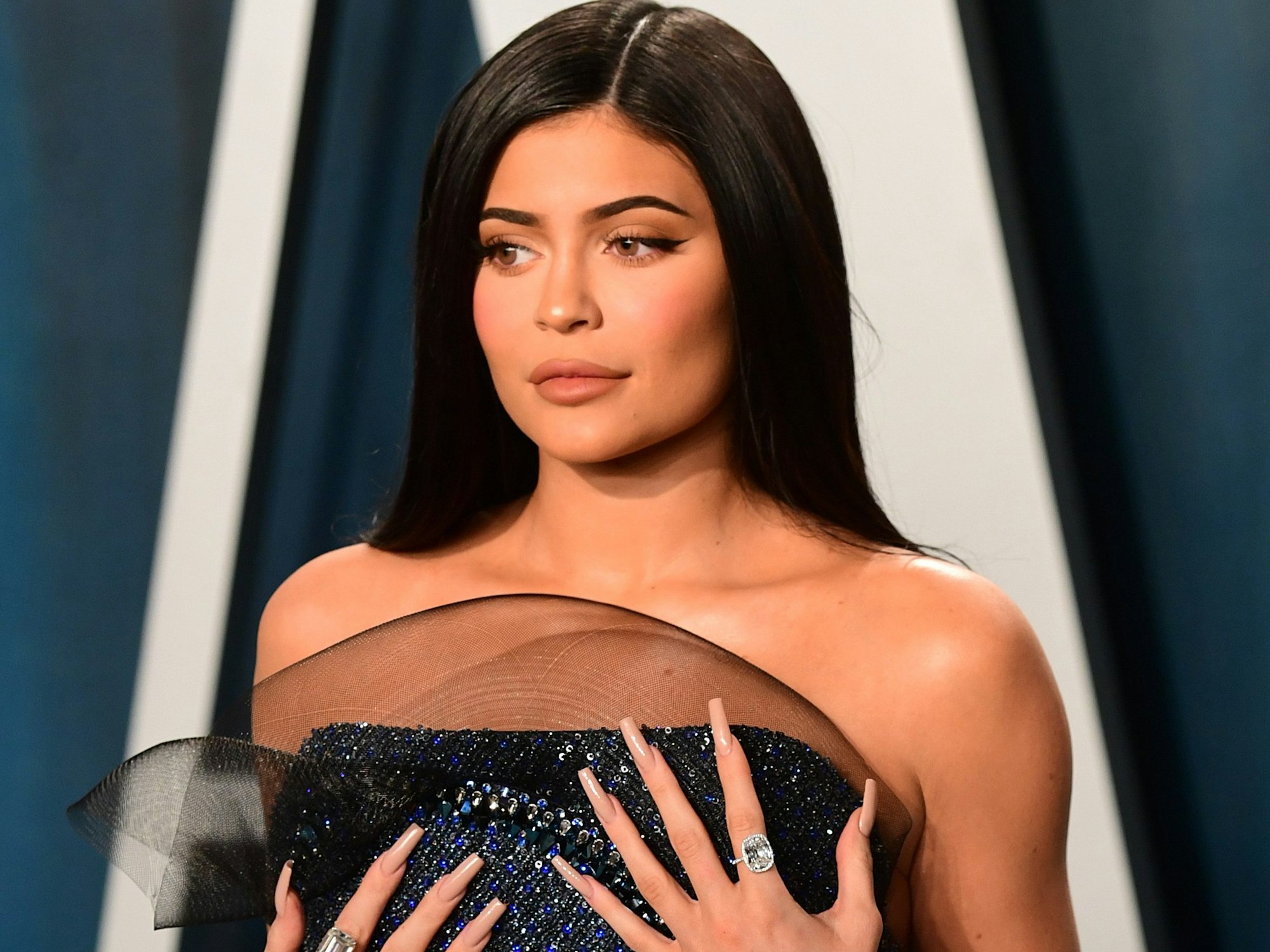 Kylie Jenner steht mit einem leichten Schmunzeln auf den Lippen vor einer blauen Wand. Die Hände hat sie auf ihre Brüste gelegt.